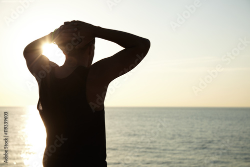Back shot of fit black footballer holding his clasped hands on his nape, contemplating wonderful landscape of sea at sunset posing against copy space background for your promotional content