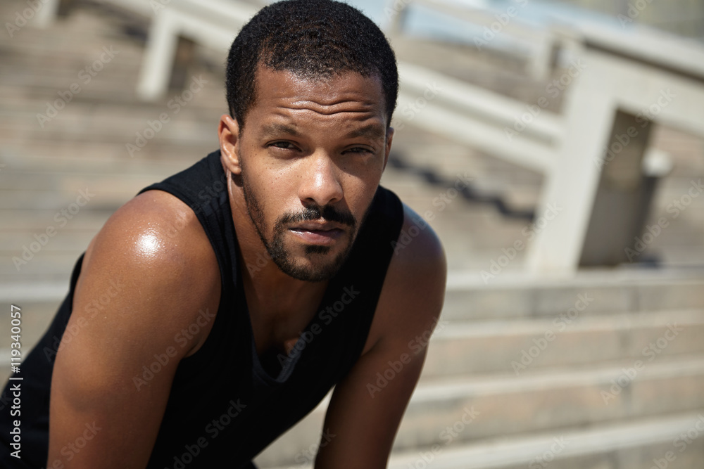 Attractive young black jogger leaning over to catch his breath, having rest during break while training outdoors, looking at camera with serious expression, squinting eyes because of bright sunshine