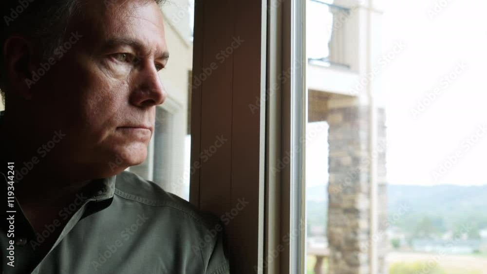 An unsmiling mature man standing at a window deep in thought about something serious.
