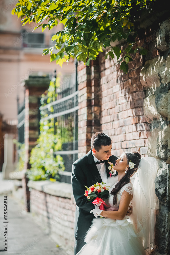 Obraz premium Handsome groom hugging beautiful bride with bouquet in romantic european park