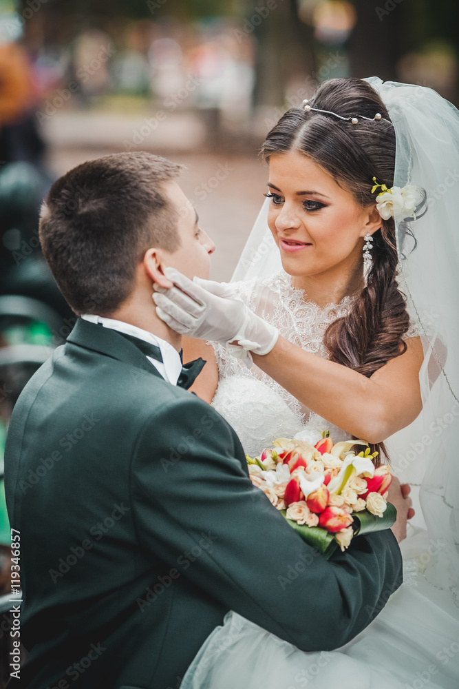 Bride and groom sitting on bench in park, holding hands of each other and bouquet. Groom holding his head on bride's shoulder and bride smiling broadly. Outdoor, full body Bride and groom sitting on bench in park, holding hands of each other and bouquet. Groom holding his head on bride's shoulder and bride smiling broadly. Outdoor, full body
