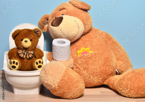 Small teddy bear sitting on the potty and playing with toilet paper, father bear teaching his cute kid how to pee and poo