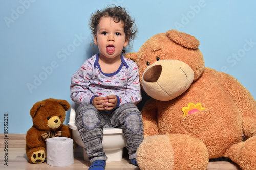 Baby toddler sitting on a potty surrounded by teddy bears. Cute kid potty training for pee and poo helped by teddy bear who gives him toilet paper