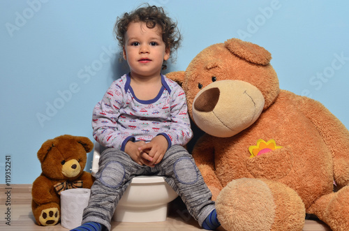 Baby toddler sitting on a potty surrounded by teddy bears. Cute kid potty training for pee and poo helped by teddy bear who gives him toilet paper