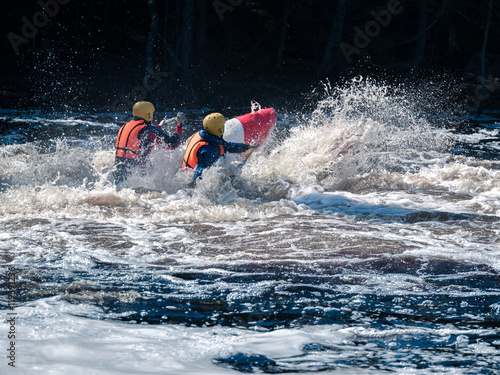 Toned image coordinated team of rowers in helmets and life jackets which sit on the raft, and fighting the rough river against the backdrop water and foam spray