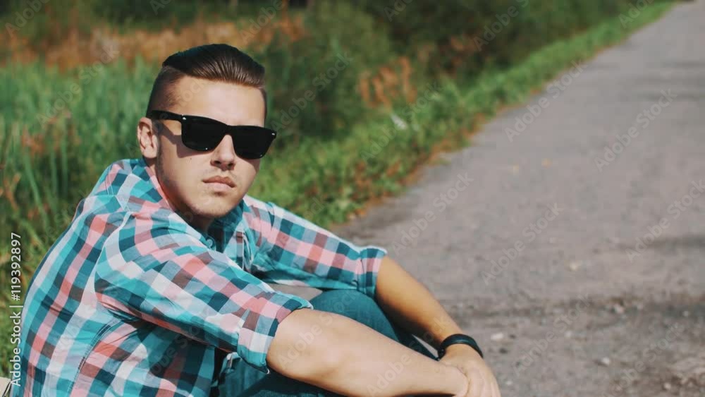 Boy sitting at road put on sunglasses. Waiting. Travel. Hitchhiking. Cars. Summer sunny day Boy sitting at road put on sunglasses. Waiting. Travel. Hitchhiking. Cars. Summer sunny day