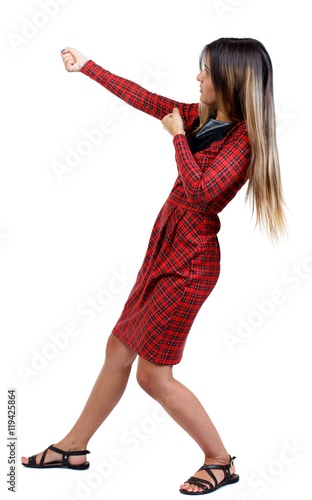 back view of standing girl pulling a rope from the top or cling to something. girl in red plaid dress stands sideways and pulling rope.
