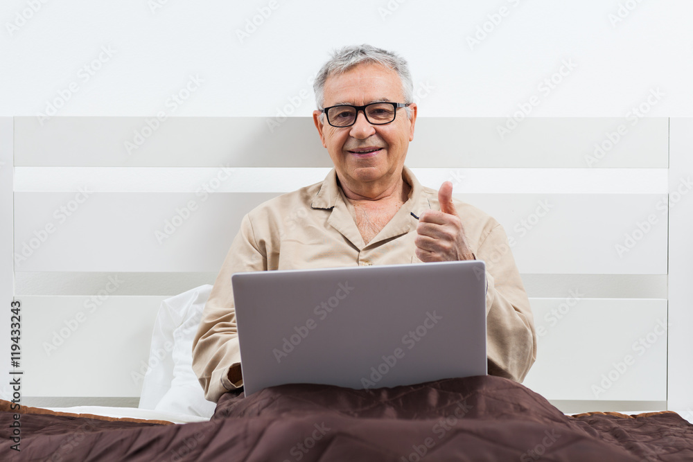 Senior man is lying in bed and using laptop.