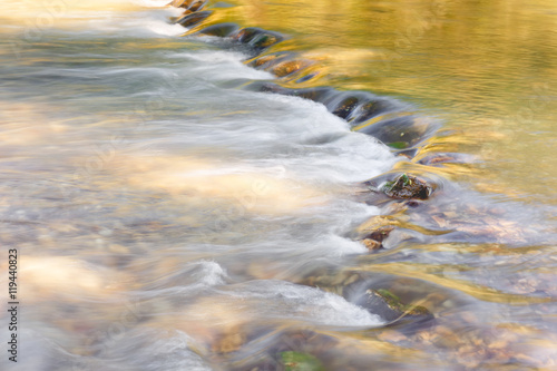 River flowing through golden and green foliage and rocks at sunlight, close up