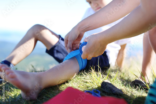Złamana noga, wypadek na górskim szlaku. Matka opatruje dziecięce kolano bandażem elastycznym.