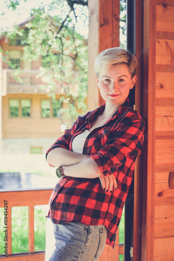 Fashionable portrait of a young beautiful woman in red lumberjack shirt, jeans, white t-shirt and sneakers. Trendy blonde hipster girl having fun on a terrace of a rich wooden house on a sunny day. Fashionable portrait of a young beautiful woman in red lumberjack shirt, jeans, white t-shirt and sneakers. Trendy blonde hipster girl having fun on a terrace of a rich wooden house on a sunny day.