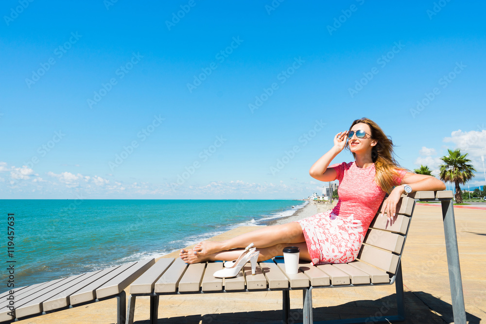 Young beautiful smiling woman in red dress sitting on beach chair with shoes, coffee and looking on blue sea and sky