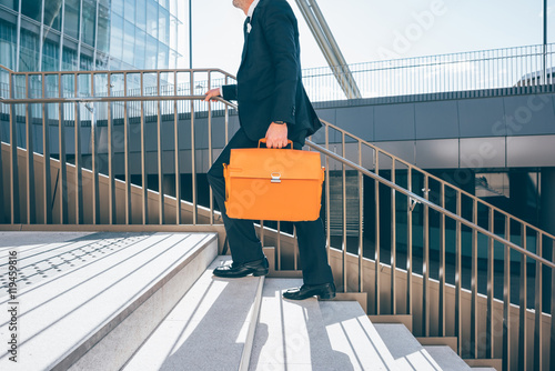 From the neck down view of a contemporary business walking upstairs in the city backlight, holding overnight bag - business, work concept