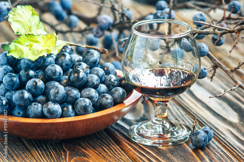 Sloe gin. Glass of blackthorn homemade light sweet reddish-brown liquid. Sloe-flavoured liqueur or wine decorated with fresh juicy ripe prunus spinosa berries on wooden background. Selective focus