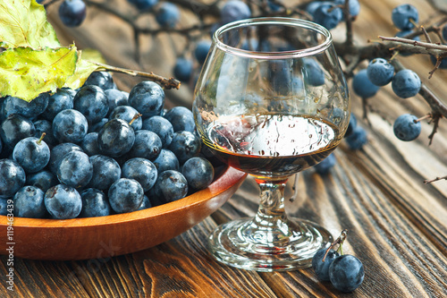 Sloe gin. Glass of blackthorn homemade light sweet reddish-brown liquid. Sloe-flavoured liqueur or wine decorated with fresh juicy ripe prunus spinosa berries on wooden background. Selective focus
