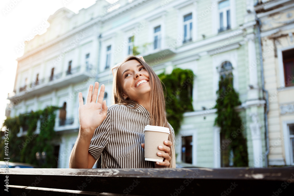 Fototapeta premium Young beautiful girl sitting on bench, holding coffee, smiling.