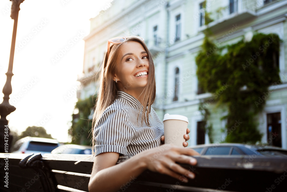 Young beautiful girl sitting on bench, holding coffee, smiling. Young beautiful girl sitting on bench, holding coffee, smiling.