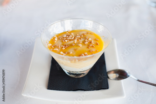 custard with chopped nut crocanti in elegant crystal glass on black paper napkin on white square plate with metal spoon on tablecloth
 photo