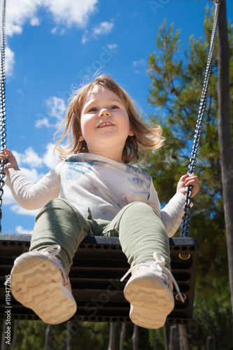 low angle shot of two years old blonde child with white shirt and green trousers teeter in swing at urban park
