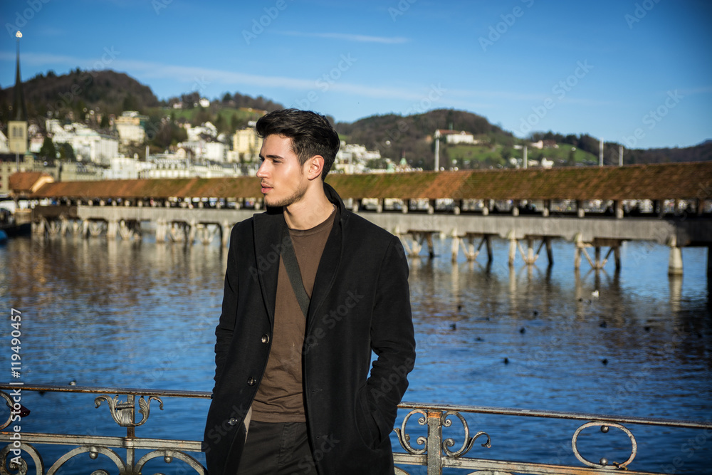 Fototapeta premium Handsome man standing near metal fence in Lausanne. Famous wooden bridge and Water Tower on background.