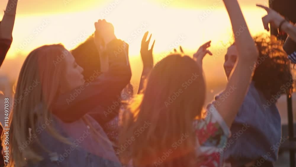 Group of young multi-ethnic people dancing with raised arms to the music played by dj at rooftop party at sunset