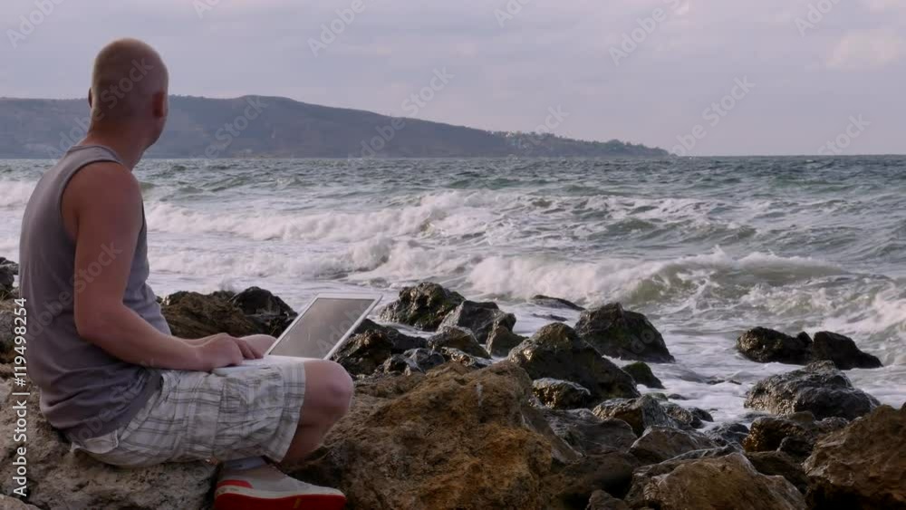 a Young Man Sitting on the Beach With a Laptop on the Rocks. the Waves Beat Against the Rocks. a Young Man Sitting on the Beach With a Laptop on the Rocks. the Waves Beat Against the Rocks.