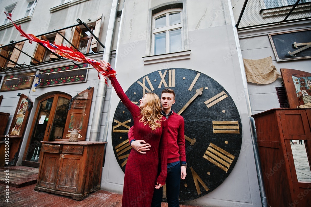 Young beautiful stylish fashion couple in a red dress in love story at the old city, play with shawl background big clock