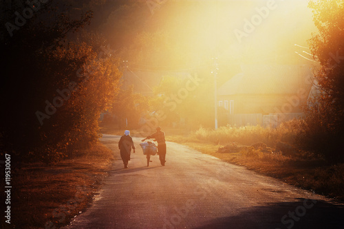 Fototapeta Naklejka Na Ścianę i Meble -  Evening walk. Elderly couple with bag on bicycle walking along road in beautiful evening light. Rural landscape with farmers. Backlight.