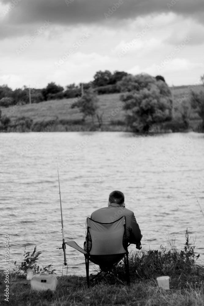 Fisherman on river. Man in folding chair on river bank expects fish. Landscape with storm clouds. Black and white photo. Fisherman on river. Man in folding chair on river bank expects fish. Landscape with storm clouds. Black and white photo.
