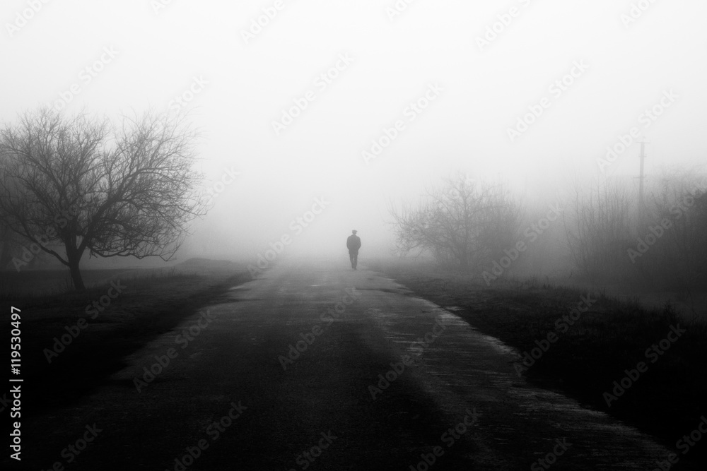 Wayfarer in fog. Silhouette of man walking on misty village road. Homecoming. Loneliness, .nostalgia, sad mood. Black and white photo - obrazy, fototapety, plakaty 