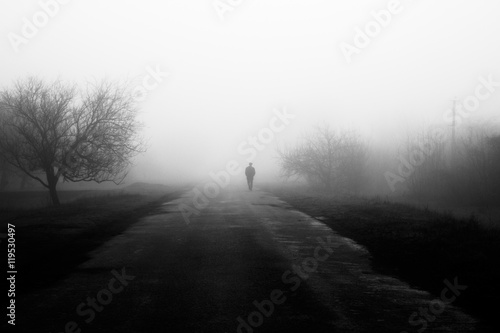 Wayfarer in fog. Silhouette of man walking on misty village road. Homecoming. Loneliness, .nostalgia, sad mood. Black and white photo photo