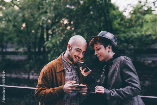 Two young caucaisan man and woma outdoor in the city using smart phone hand hold, talking and smiling - technology, social network, freindship concept photo