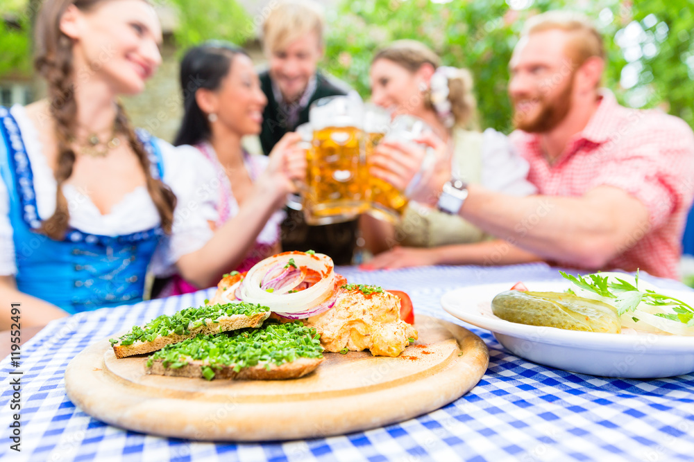 Fünf Freunde, Männer und Frauen, stoßen im Biergarten mit Bier an, bayerische Brotzeit steht auf dem Tisch Fünf Freunde, Männer und Frauen, stoßen im Biergarten mit Bier an, bayerische Brotzeit steht auf dem Tisch