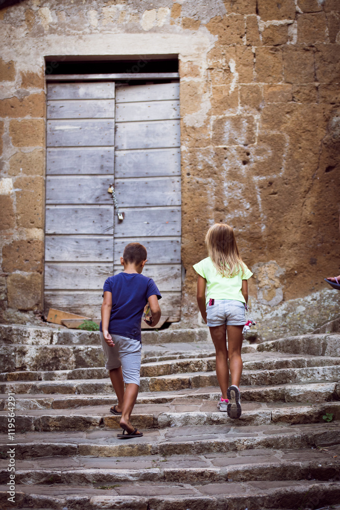 un couple d'enfant de dos, montent ensemble les marches d'un vieille escalier, avec une grande porte en haut