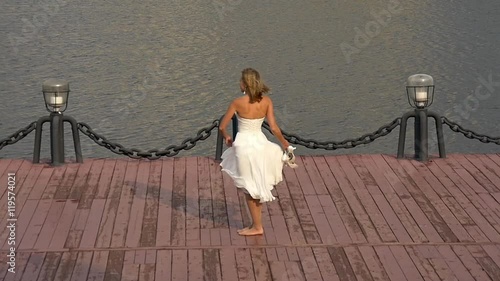 Joyful beautiful bride spinning in dance on dock near glaring water, slow motion photo