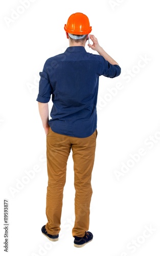 Backview of business man in construction helmet stands and enjoys tablet or using a mobile phone. man in a blue shirt and a helmet talking on the phone with his hand in his pocket.
