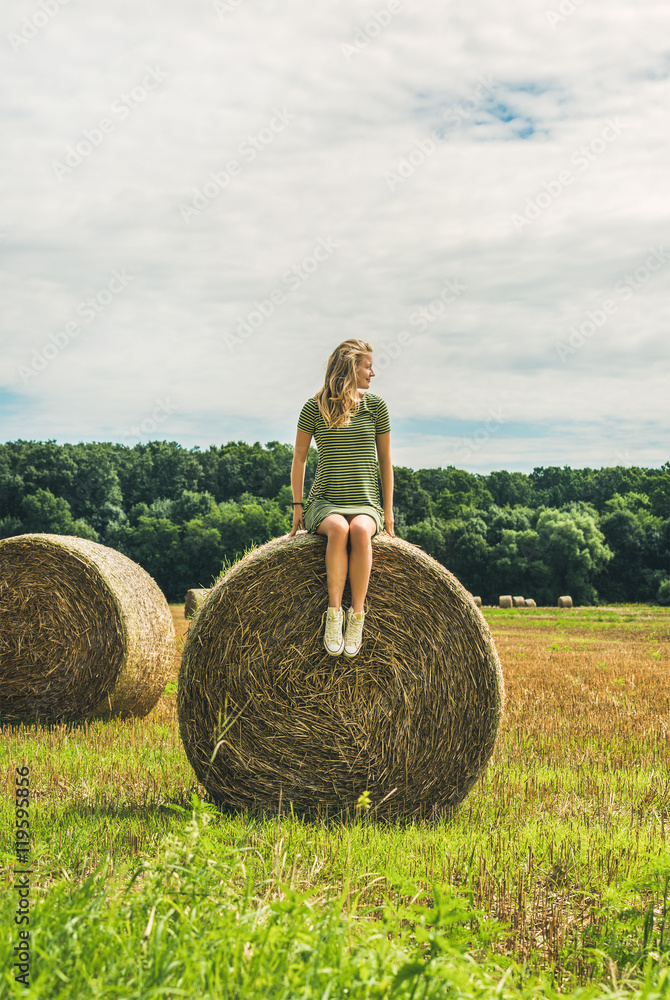Young blond lady sitting on haystack and looking away on sunny summer day, Badasconytomaj, Hungary Young blond lady sitting on haystack and looking away on sunny summer day, Badasconytomaj, Hungary