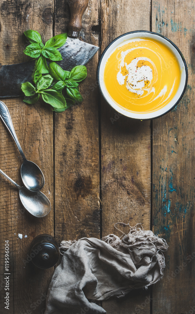 Pumpkin cream soup in bowl with fresh basil and spices over old rustic wooden background, top view, copy space, vertical composition