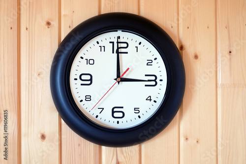 Big round wall clock with a black rim with arrows showing three o'clock hangs on brown wooden wall from vertical planks horizontal view indoor close-up