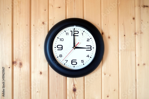 Big round wall clock with a black rim with arrows showing three o'clock hangs on brown wooden wall from vertical planks horizontal view indoor on wooden background