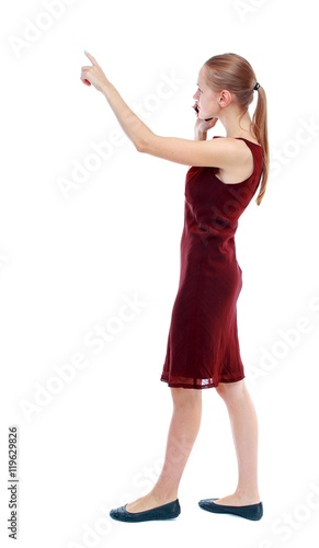 back view of pointing young beautiful girl with tablet computer. girl watching. Isolated over white background. The girl in the maroon sleeveless dress goes sideways and talking on a smartphone and