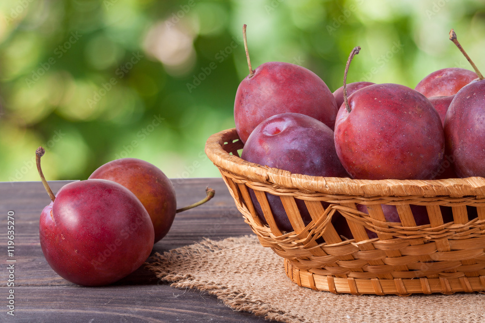 plum in a wicker basket on the wooden table with sackcloth and blurred green background plum in a wicker basket on the wooden table with sackcloth and blurred green background