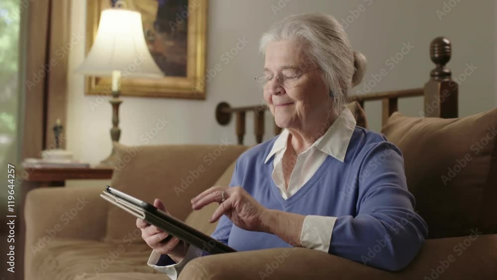 A smiling senior elderly woman uses a computer tablet device on her couch. Shot in 4K UHD.