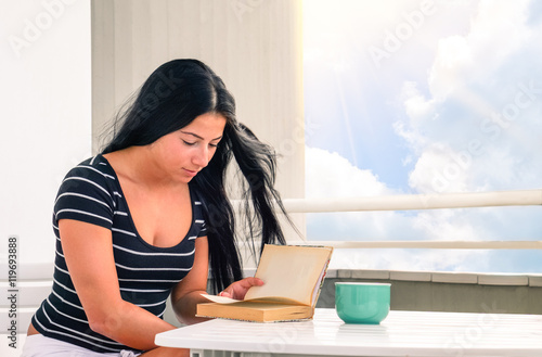 Young woman reading book with tea cup on table - Beautiful girl studying at home sitting on terrace with white clouds sky as background - Concept of female relax in peaceful and candid ambience 