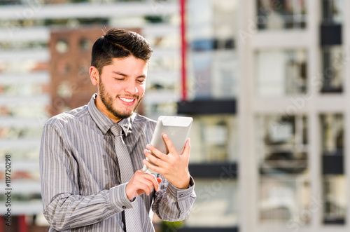 Young handsome man wearing shirt and tie standing on rooftop, holding tablet staring at screen, city buildings background photo