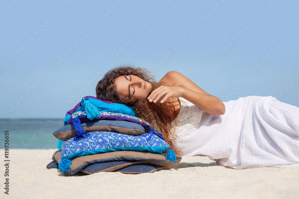 Beautiful woman in white dress lying on pillows at the beach