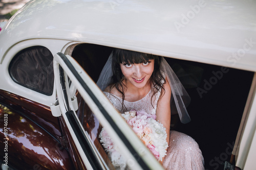 Gorgeous stylish brunette bride posing in retro brown and white limousine car in white dress. Woman near vehicle