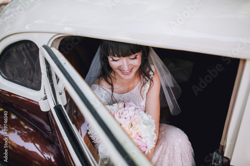 Gorgeous stylish brunette bride posing in retro brown and white limousine car in white dress. Woman near vehicle
