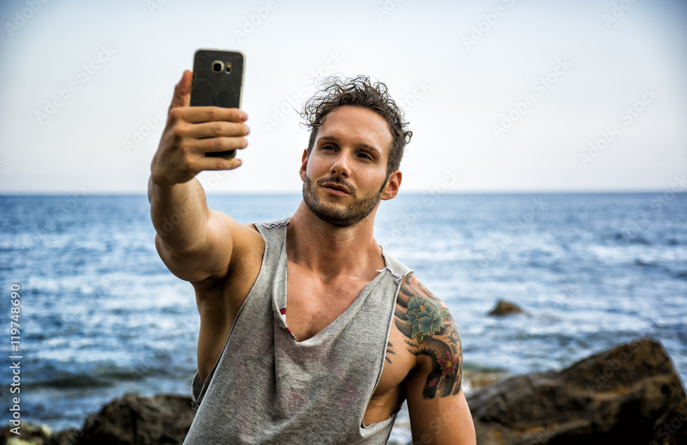 Athletic man at the seaside using cell phone to take selfie photo with the sea behind him
