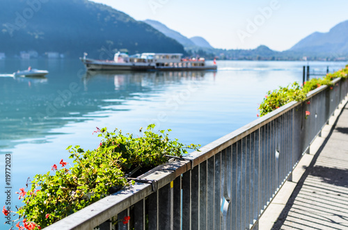 Vacation summer sunny lake view. Beautiful flowers over the background of beautiful serene blue Lugano lake, clear blue sky and a ship surrounded by hills in Morcote, Switzerland, selective focus